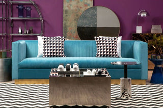 A modern living room featuring the Premier Sofas Contemporary Light Blue Velvet 3 Seater Sofa with gold base and tufted design, paired with geometric pillows, a metallic coffee table, zigzag rug, abstract art, and a round mirror on a purple wall.