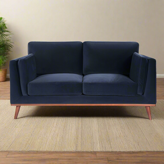The Maili Velvet Fabric 2 Seater Sofa by Twenty Ten Designs in midnight blue offers cushioned comfort and wooden legs. It sits on a beige rug against a light wall, with a green potted plant to the left in the background.