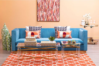 A stylish living room with the Premier Sofas Contemporary Light Blue Velvet 3 Seater Sofa (gold base, tufted), geometric pillows, glass coffee table, orange rug, abstract art, green jar, and a gold bar cart with flowers and drinks.