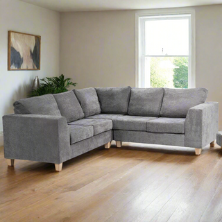 A modern living room showcases the Budget Sofa Collections SOFAINSTORE Grey Textured Fabric Corner Sofa with wooden legs on light hardwood floors, complemented by a potted plant, natural light from a window, and abstract art on a beige wall.