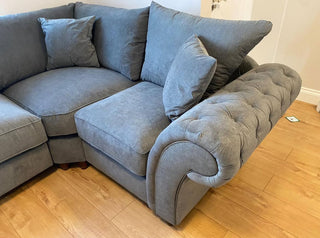 The Beehive Furniture Windsor Fullback Grey Corner Sofa | Right Hand Facing, featuring plush cushions and a tufted armrest, is placed on a light wooden floor in a modern living room with a small white object nearby.