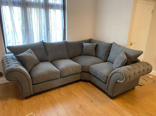 The Beehive Furniture Windsor Fullback Grey Corner Sofa | Right Hand Facing with plush cushions and rolled arms is set in a modern living room with light walls, wooden floor, and a window with sheer curtains.