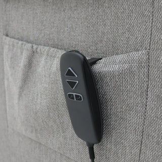 A black remote control with arrow buttons rests in the side pocket of the RS Furniture SOFAINSTORE Bramley Grey Electric Rise & Recline Armchair – a light grey dual motor comfort lift chair upholstered in soft fabric.