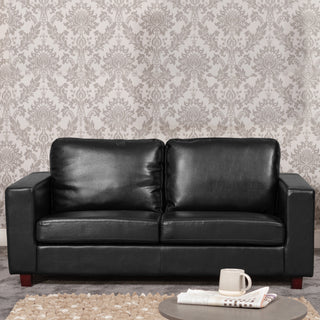 The KH Sofas Woodleigh Leather 3 Seater Sofa in black with comfortable foam cushions sits before patterned wallpaper, while a coffee table with a white mug and newspapers stands on a beige rug in the foreground.