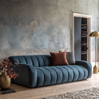 A Gallery Direct Coste Fabric 3 Seater Sofa in blue upholstery, featuring retro design and comfort, sits on a wooden floor with two throw pillows, beside a gold lamp and side table with a vase in front of a textured blue-gray wall.