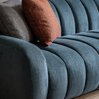 A close-up of Gallery Direct’s Coste Fabric 3 Seater Sofa in blue upholstery with retro-inspired vertical channeling, shown with a textured brown pillow and an orange pillow.