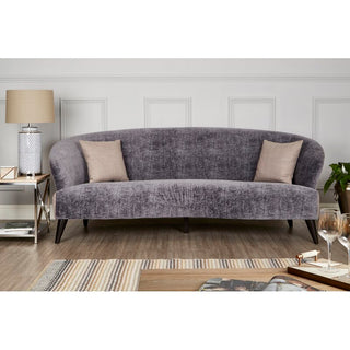 A modern living room showcases the Premier Sofas Contemporary 3 Seater Sofa in grey velvet with slim black legs, styled with two beige pillows, a side table holding a lamp, books, and decor against white paneled walls and hardwood floors.