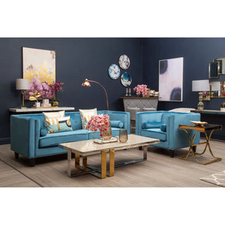A modern living room with blue walls, a Premier Sofas Felisen Upholstered Velvet 3 Seater Sofa in Blue Elegance, a marble coffee table, floral arrangements, wall art, decorative clocks, gold accent furniture, and light wood floors.