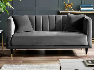 The Julian Bowen SOFAINSTORE Savona Grey Velvet 2 Seater Sofa features a channel-tufted back, gold-tipped legs, and comes with two matching pillows—ideal for luxe modern UK interiors. Displayed with chic decor against a dark paneled wall.