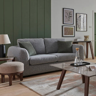 A modern living room featuring the Vida Living Santiago Fabric 2 Seater Sofa with grey upholstery and black legs, two green cushions, a round beige ottoman, wooden lamp, marble coffee table set, stylish wall art, and green accent wall panels.