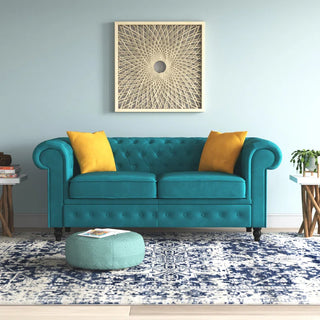 The PF Sofas SOFAINSTORE Florencia Ocean Teal Velvet Chesterfield 2 Seater, with scroll arms and button tufting, stands against a light blue wall under geometric art. A patterned rug, teal ottoman, side tables, and a plant finish the modern room.