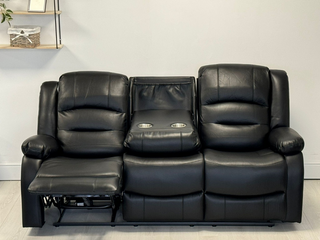 The Beehive Furniture SOFAINSTORE Arclay Black Leather Recliner Sofa is a 3-seater with electric recline, a drop-down cupholder console, and the left footrest extended. It sits against a light wall with a small shelf in the background.