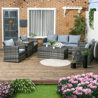 A cozy patio boasts the Outsunny 6 Pieces Rattan Garden Furniture Set, a 7-seater grey wicker sofa set with cushions, storage tea table, and side table, surrounded by potted plants, string lights, and pink flowers against a brick wall.