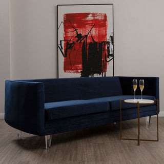 A modern living room showcases the Premier Sofas Modern Dark Blue Velvet 3 Seater Sofa with sleek acrylic legs, a chic side table with champagne glasses, and an abstract red and black painting on the wall behind.