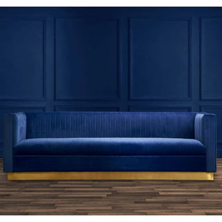 The Premier Sofas Deep Blue Velvet 3 Seater Sofa with a gold base and elegant design is set against a matching dark blue paneled wall on a wooden floor.