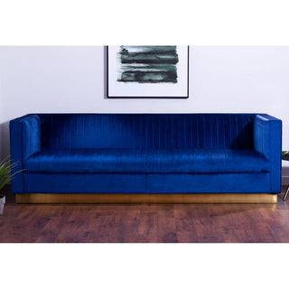 The Premier Sofas Deep Blue Velvet 3 Seater Sofa, with a gold base and elegant design, is set against a white wall under an abstract black-and-green painting. Wooden floors and a small plant complete the chic space.