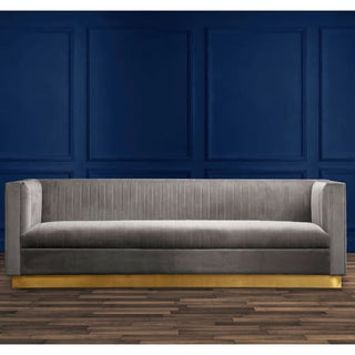 The Premier Sofas Grey Velvet 3 Seater Sofa with a gold base and channel tufting sits in front of a dark blue paneled wall on a wooden floor.