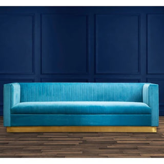 The Premier Sofas Contemporary Light Blue Velvet 3 Seater Sofa with a gold base and tufted design sits against a dark blue paneled wall on a wooden floor.