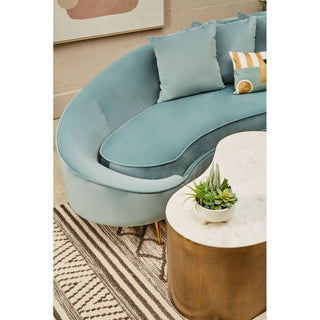 A modern living room features the Premier Sofas Osmodin 4 Seater Curved Sofa in light blue velvet, paired with a white marble-top coffee table with a gold base, a small potted succulent, and a geometric-patterned striped rug.
