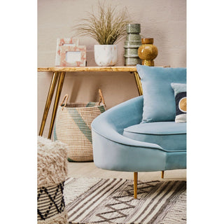 A modern living room with the Premier Sofas Osmodin 4 Seater Curved Sofa in light blue velvet, accessorized with geometric cushions, a patterned rug, wooden console table, woven basket, potted plant, and a neutral wall backdrop.