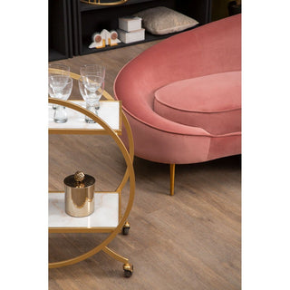 A modern living room features the Crescent Salmon Pink Velvet Sofa by Premier Sofas, a curved 4 seater with luxurious design, plus a round gold and white bar cart with three glasses and a gold ice bucket on wooden flooring.