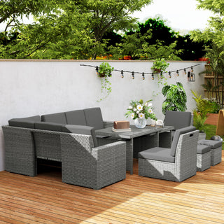 A modern patio is styled with the Outsunny Seven-Piece Rattan Garden Sofa Set in light grey, featuring sofas, armchairs, and an expanding glass-top table on a wooden deck. Green plants, flowers, and string lights enhance the look against a white wall.