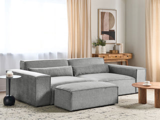 A modern living room with the Beliani SOFAINSTORE Grey Fabric 4 Seater Sofa—Oversized, deep cushions, wide armrests—a matching ottoman, wooden console table with vase, abstract wall art, beige curtains, round coffee table, side table, and patterned rug.